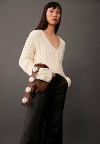 Kremfarvet kabelstrik cardigan med dyb V-hals, parret med sorte brede bukser og en brun clutch med metalknapdetaljer.