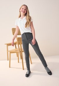 Polo blanc à manches courtes, pantalon gris ajusté avec poches avant, ballerines noires et chaussettes grises, positionnés près d'une chaise en bois.