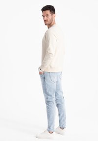 Felpa beige con collo a giro, abbinata a jeans in denim azzurro chiaro e sneaker bianche. Modello in piedi di spalle, con le mani in tasca.