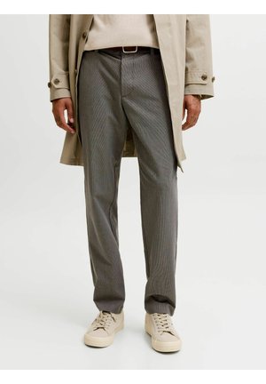 Homme portant un pantalon gris à carreaux, un manteau beige et des baskets beiges, debout sur un fond clair uni.