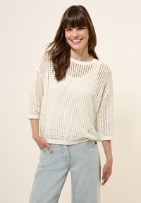 Leichter, weißer Strickpullover mit offenen Maschen, Rundhalsausschnitt und Dreiviertelärmeln, kombiniert mit hellblauen Jeans.