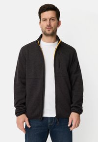 Schwarze Fleecejacke mit Reißverschluss, hohem Kragen, orangem Futter und Brusttasche. Über einem weißen T-Shirt getragen, kombiniert mit blauen Jeans.