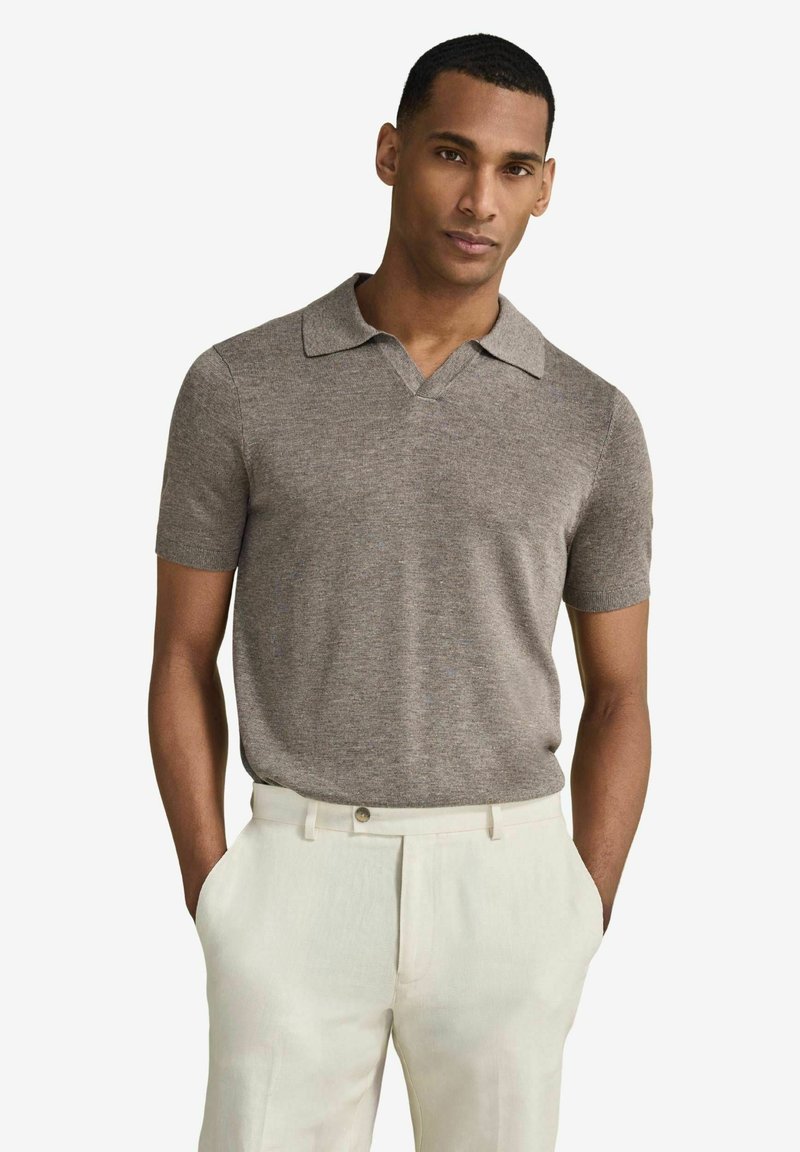 Homme portant un polo gris en maille à manches courtes rentré dans un pantalon blanc taille haute, les mains dans les poches, debout devant un fond uni.
