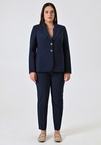 Marineblauwe op maat gemaakte pak met een eenrijige blazer met zilveren knopen, aangevuld met slanke broek en beige loafers.