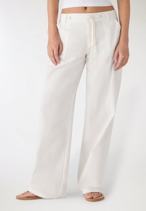 Donna che indossa pantaloni bianchi a gamba larga con coulisse e infradito beige, in piedi con le mani rilassate lungo i fianchi, su sfondo semplice.