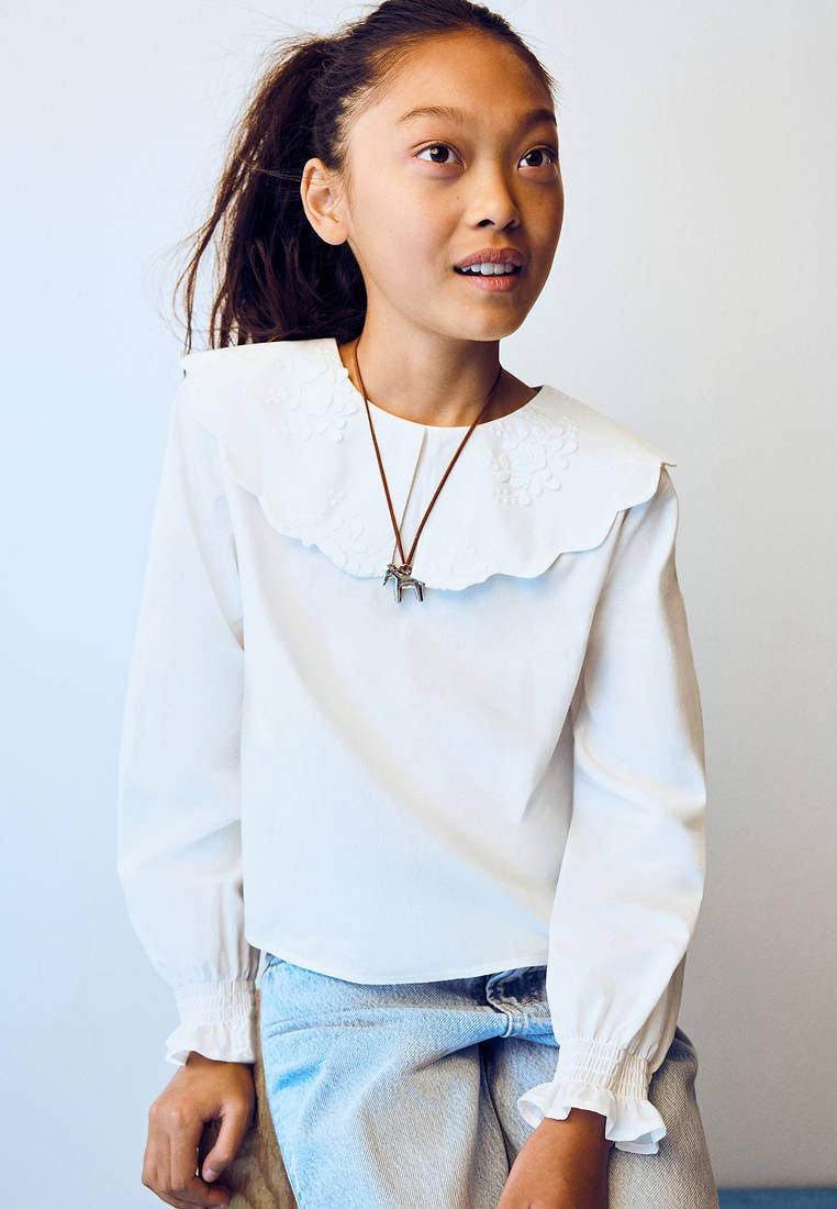 Fille avec une queue de cheval portant un chemisier blanc avec un grand col brodé et un collier pendentif cheval, assise et regardant sur le côté.