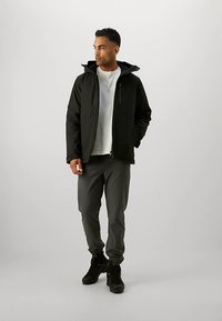 Uomo in piedi con una giacca a vento nera con cappuccio, una maglietta bianca, pantaloni grigi e stivali da escursionismo neri, che guarda verso sinistra.