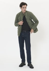 Ein Mann trägt eine steppte olivgrüne Jacke über einem schwarzen Hemd, dunkle Jeans und schwarze Wanderstiefel und steht vor einem weißen Hintergrund.