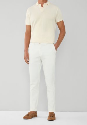 Modèle masculin portant un polo crème à manches courtes, un pantalon blanc ajusté et des mocassins à pampilles en daim marron, debout contre un mur gris uni.