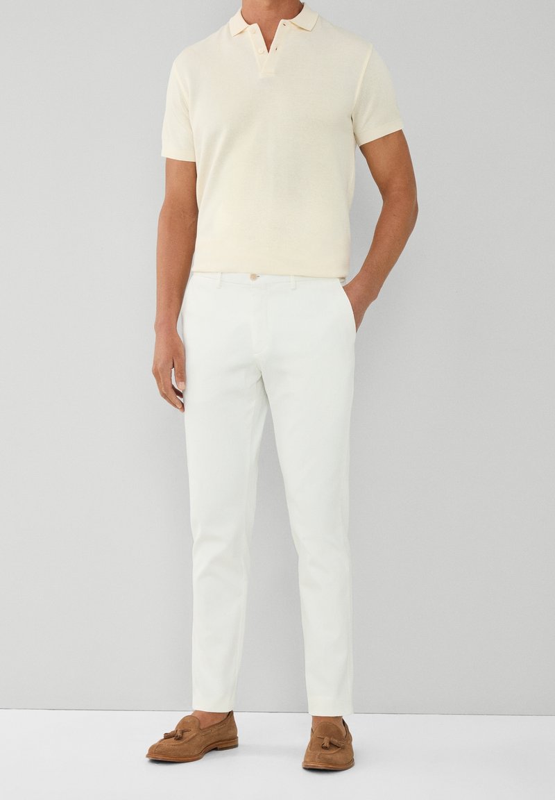 Modèle masculin portant un polo crème à manches courtes, un pantalon blanc ajusté et des mocassins à pampilles en daim marron, debout contre un mur gris uni.