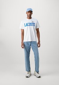 Balta medvilnė marškinėliai su mėlynu "LACOSTE" logotipu, derinami su šviesiai mėlynomis džinsomis ir pilkais sportiniais bateliais su geltonais akcentais. Šviesiai mėlyna kepurė.