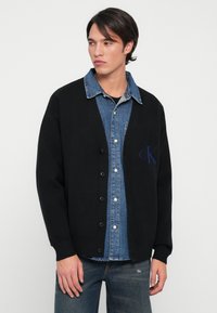 Cardigã preto feito de material suave, apresentando um decote em V, fechamento com botões e o logótipo da cK. Usado sobre uma camisa de ganga com colarinho e lavagem clara.