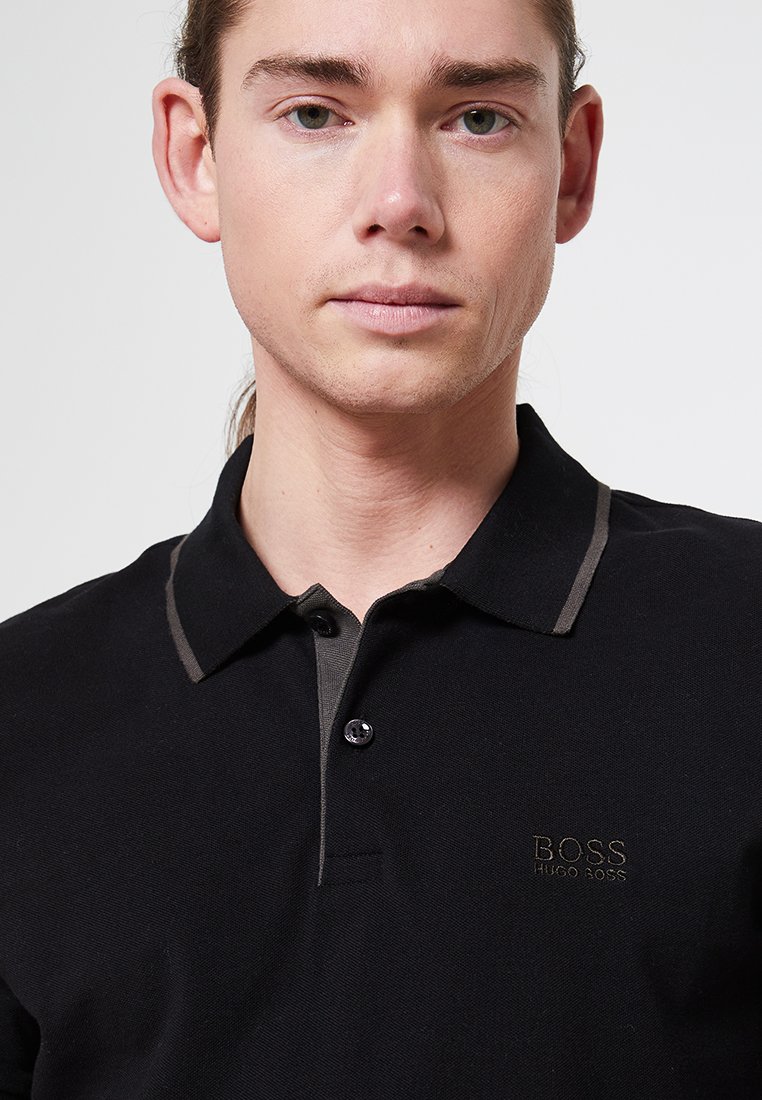 Polo-shirt noir avec un col bicolore, présentant un logo brodé "BOSS HUGO BOSS." Texture lisse et fermeture à boutons sur le devant.