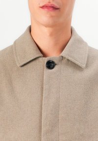 Beige wollen jasje met een kraaghalslijn, een enkele zwarte knop en een gladde, gestructureerde stof. Close-up van de bovenkant van de voorkant.