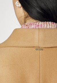 Vue arrière d'une personne portant un manteau camel avec une épingle dorée au logo "BOSS" et un col tricoté multicolore visible.