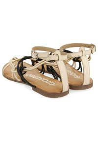 Beige a černé ploché sandály na řemínky s přezkami kolem kotníků a hnědými stélkami označenými 'GIOSSERPO' na bílém pozadí.