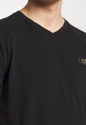 Camiseta negra de pico con un ajuste slim, mangas raglán cortas y un acento de logotipo dorado en el área del pecho izquierdo. Textura de tela suave.
