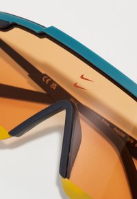 Akiniai nuo saulės su dideliu, apvaliu oranžiniu lęšiu, melsva viršutine juosta ir geltonais akcentais. Juodi smailėjantys rėmeliai su Nike logotipo detalėmis.