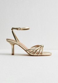 Guldklädda sandaler med remmar och en vriden framdesign, ankelrem och en smal, metallisk stilettklack. Slät yta och öppen tå.
