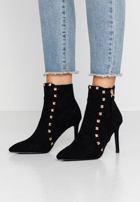 Bottes courtes en daim noir avec bout pointu, fermeture éclair latérale et clous métalliques dorés sur le devant. Talon stiletto haut. Jeans bleus effilochés visibles.