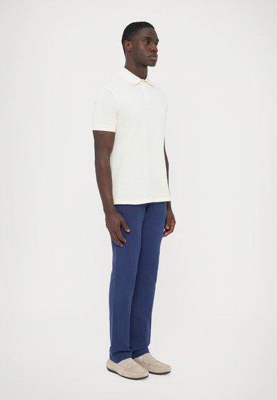 Krémové polo tričko s textúrovaným vzorom, skombinované s tmavomodrými nohavicami a svetlobéžovými mokasínami. Strih je prispôsobený a moderný.