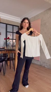 Νεαρή γυναίκα με σκούρα μαλλιά κρατάει ένα λευκό μακρυμάνικο τοπ σε μια κρεμάστρα σε ένα μοντέρνο, λιτά διακοσμημένο δωμάτιο με ξύλινο πάτωμα.