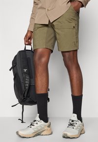 Shorts cargo color khaki con tasche, zaino nero testurizzato, scarpe da ginnastica bianche e beige, e calzini neri al polpaccio. Dettagli di design minimalista.