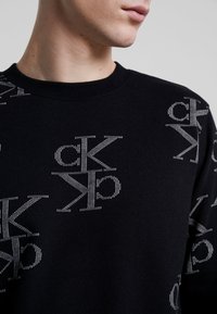 Felpa nera con un motivo a ripetizione di lettere stilizzate in argento. Realizzata in tessuto morbido con colletto e polsini a coste.