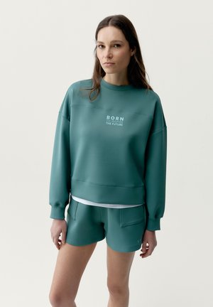 Kvinde med langt brunt hår iført teal sweatshirt og matchende shorts, sweatshirtens tekst lyder "FØDT TIL AT INSPIRERE FREMTIDEN" på ensfarvet baggrund.