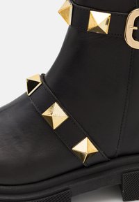 Botină neagră din piele cu gleznă, având o curea lată cu ținte piramidale aurii și o talpă din cauciuc texturat. Design durabil și modern.