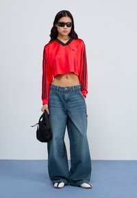 Rotes, gekürztes langärmliges Shirt mit schwarzen Streifen, blaue weite Jeans und weiße Rutschsandalen. Hält eine kleine schwarze Tasche. Glatte Textur.