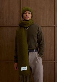 Bonnet en tricot vert et écharpe assortie en matière douce, associés à un long pull en laine marron et un pantalon gris, mettant en valeur une étiquette.