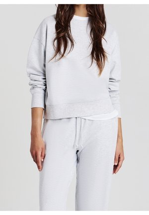 Woman wearing light grey long-sleeve sweatshirt and matching sweatpants with drawstring, standing against plain light background.
