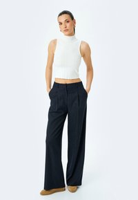 White ribbed sleeveless crop top paired with navy pinstriped wide-leg trousers and tan suede shoes. Model poses with hands in pockets.