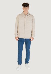 Young man with curly hair wearing a beige buttoned shirt, blue jeans, and white sneakers standing against a plain background.