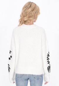Biały, oversize sweter z czarnym haftem kwiatowym na rękawach. Miękka, strukturalna tkanina, ściągacze i dół wykończone ściągaczem. W zestawieniu z jasnoniebieskimi dżinsami.