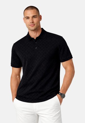 Άνδρας που φοράει μαύρο πουκάμισο πόλο με σχέδια και λευκό παντελόνι, με τα χέρια στις τσέπες και μαύρο ρολόι χειρός στο αριστερό χέρι.