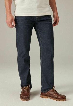 Dark blue jeans made of denim, straight-leg design, featuring yellow stitching and front pockets, paired with brown lace-up shoes.