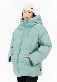 Mujer joven con una chaqueta acolchada de color verde claro con capucha y un gorro de lana blanco, con las manos en los bolsillos de la chaqueta, mirando al frente.