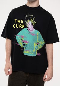 Black oversized t-shirt featuring a colorful graphic of a figure in teal and pink, with "THE CURE" text in yellow above the illustration.