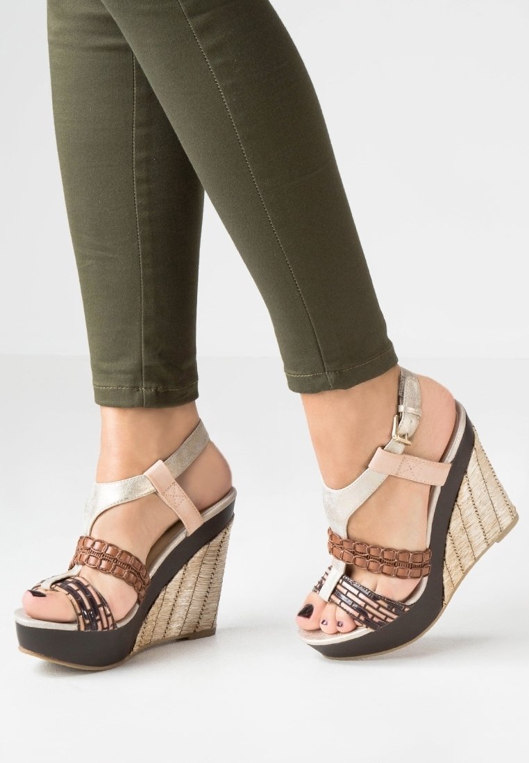 Pieds portant des sandales compensées tissées beige et marron avec des brides à la cheville et un pantalon vert ajusté.