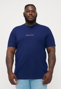 Camiseta de manga corta azul marino hecha de una tela suave, con "Originals Studio" bordado en contraste en el pecho.