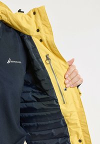 Veste imperméable jaune avec un intérieur en contraste marine, dotée d'une poche zippée, de boutons pression et d'une texture matelassée.