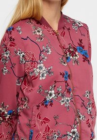 Veste bomber florale en tissu rose pâle, ornée de fleurs blanches, bleues et rouges, col côtelé et fermeture éclair dorée.