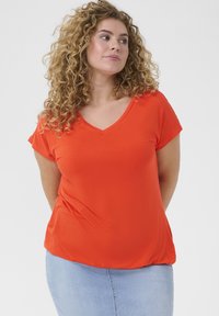 Camiseta naranja de cuello en V y mangas cortas, hecha de tela suave, con un corte relajado y un ligero caído, combinada con una falda de mezclilla azul clara.