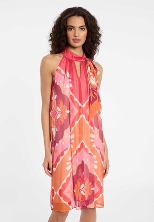 Vrouw met krullend donker haar, gekleed in een mouwloze halterjurk met roze en oranje patroon tot op de knieën, staand tegen een witte achtergrond.