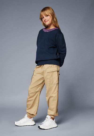 Marinblå sweatshirt med lila nackdetalj, beige cargobyxor med fickor och vita sportskor. Modellen står i en avslappnad pose.