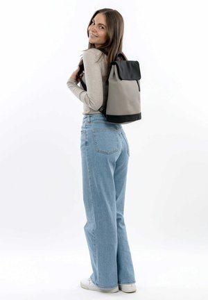 Zaino in tessuto grigio con pattina e base nera. Presenta spallacci regolabili e una zip decorativa. Il modello indossa jeans azzurri chiari.