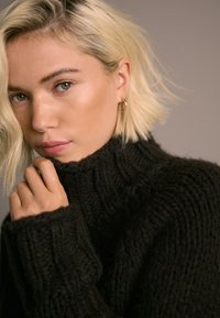 Maglione nero a collo alto in maglia con trama a coste e maniche lunghe. Il modello tiene il colletto con le mani, mettendo in risalto la morbidezza del capo.