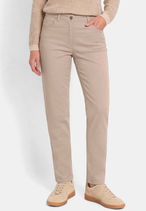 Pantalon beige droit porté avec un haut beige à manches longues et des baskets beige assorties avec semelles en gomme. Une main dans la poche, posture debout.